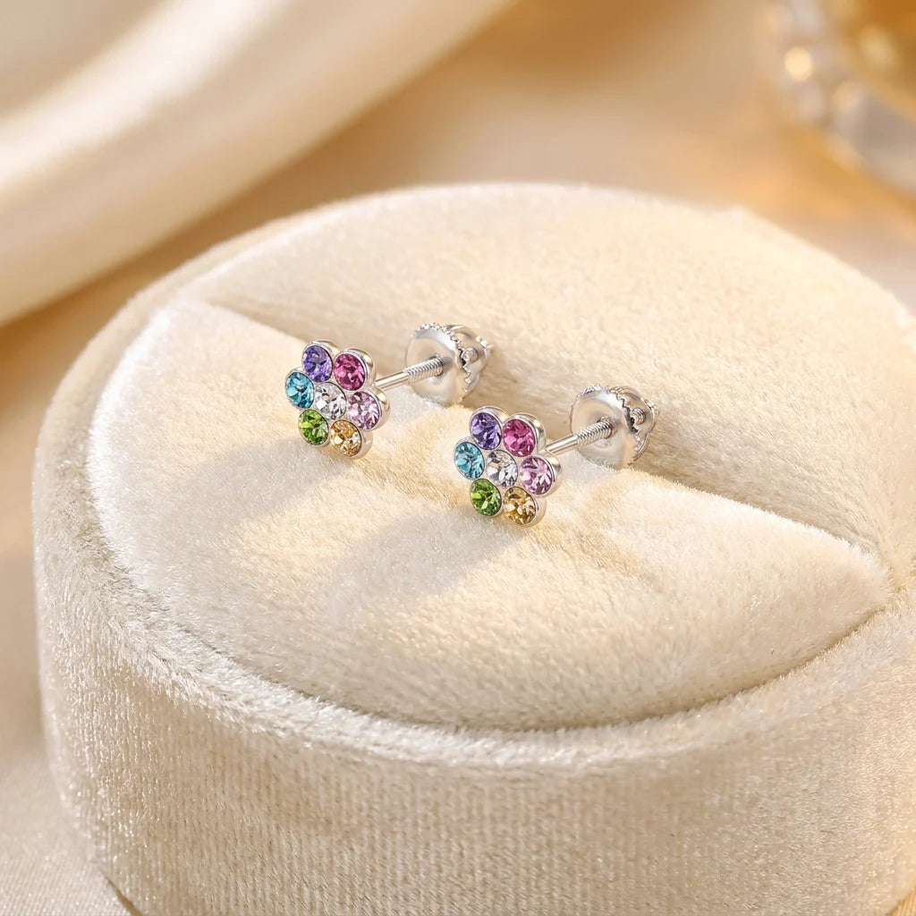 A pair of silver stud earrings with small flower-shaped clusters of multicolored rhinestones, displayed on a round cream velvet jewelry box.