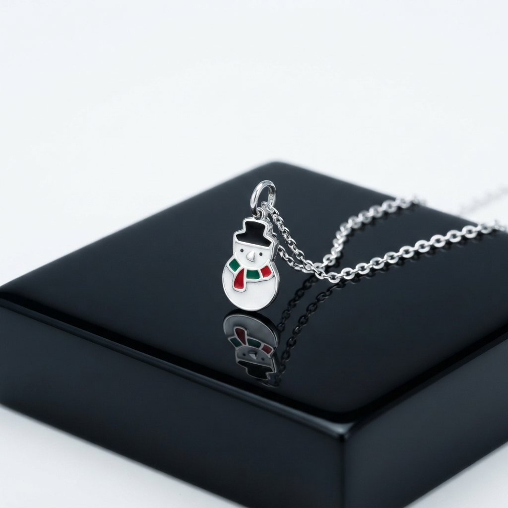 A silver snowman pendant necklace with a black hat, white body, and red and green scarf, displayed on a glossy black jewelry box against a white background.