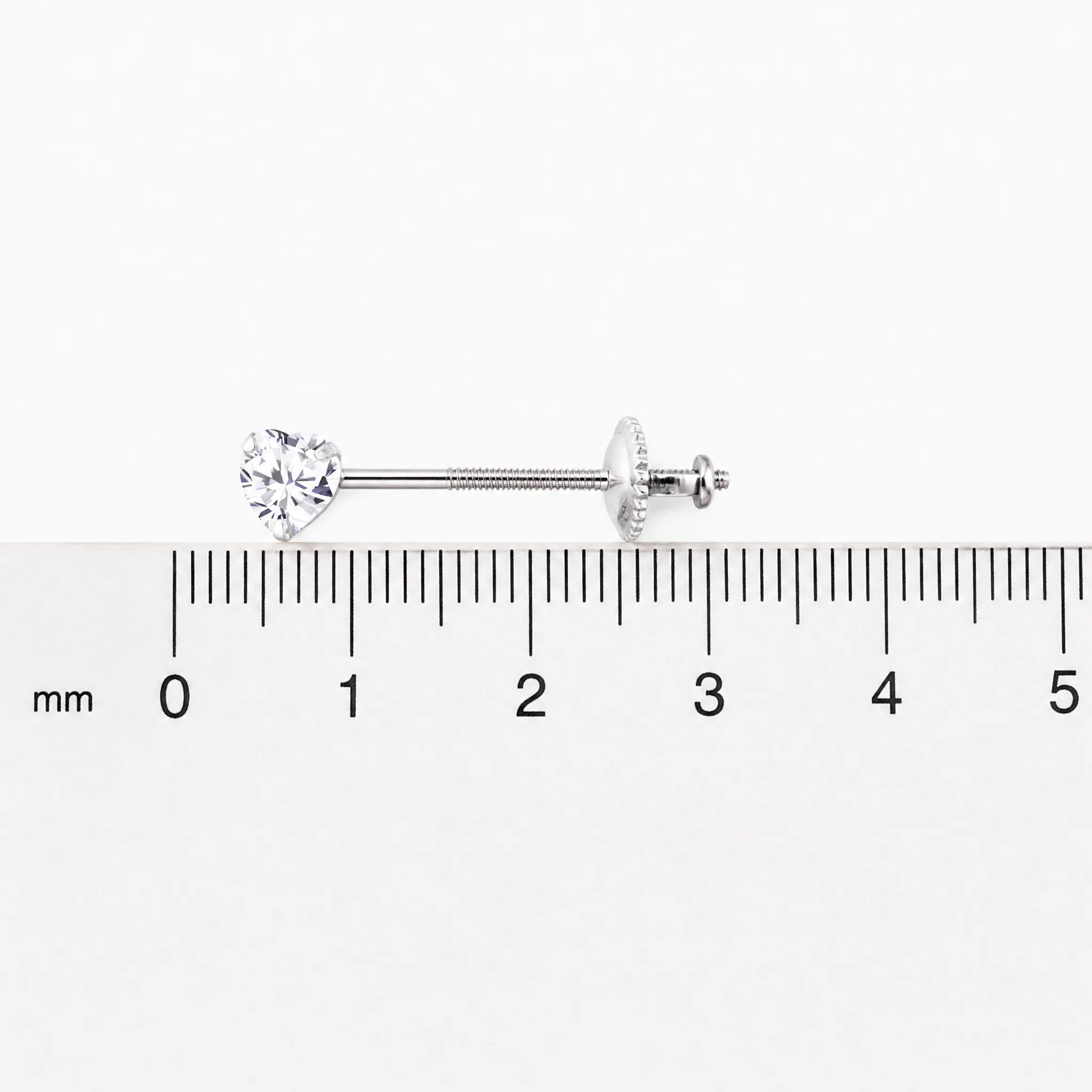 A silver screw back stud with a clear heart-shaped gemstone, shown horizontally above a millimeter ruler for scale on a white background.