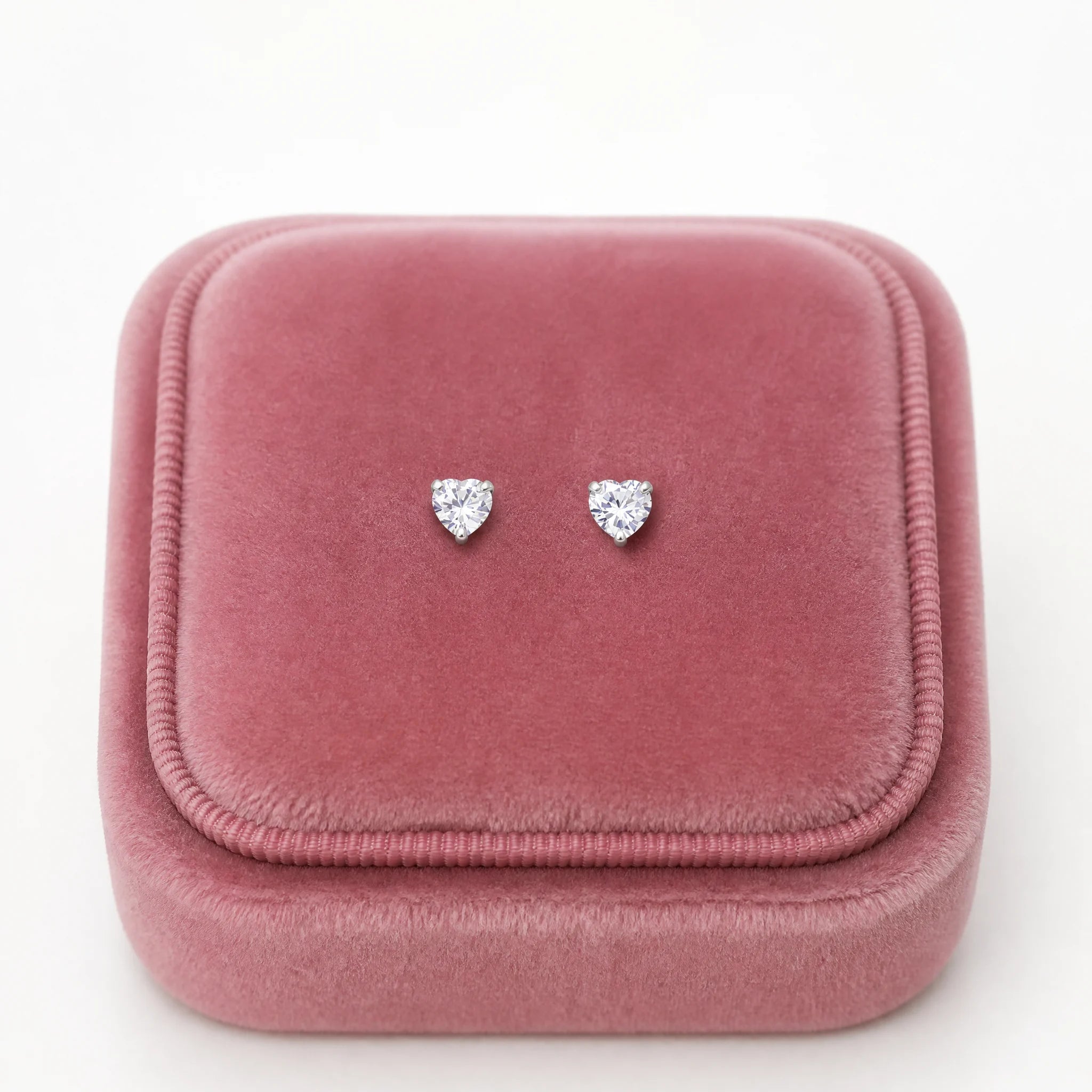 A pair of small heart-shaped stud earrings with clear sparkling stones, displayed on a dusty pink velvet jewelry box lid against a white background.