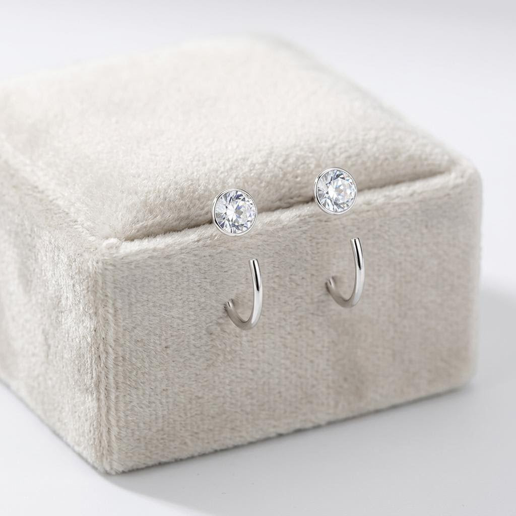A pair of silver pull through hoop earrings with round clear gemstone studs, displayed on a beige velvet jewelry box against a white background.