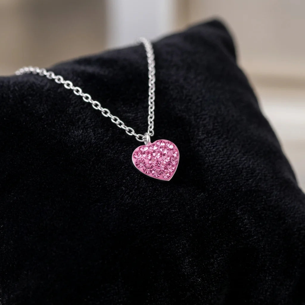 A silver chain necklace with a heart-shaped pendant covered in sparkling pink crystals, displayed on a black velvet surface.