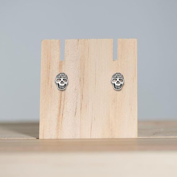 A pair of silver skull stud earrings displayed on a small wooden stand against a soft light gray background.