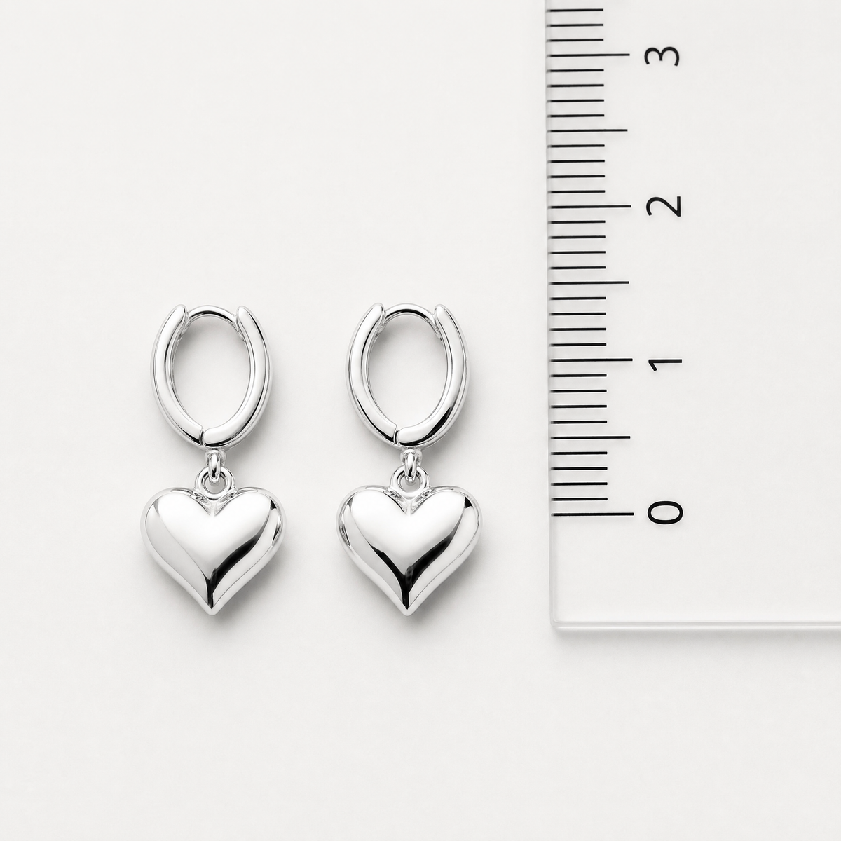 A pair of polished silver hoop earrings with dangling heart charms, displayed next to a white ruler for scale