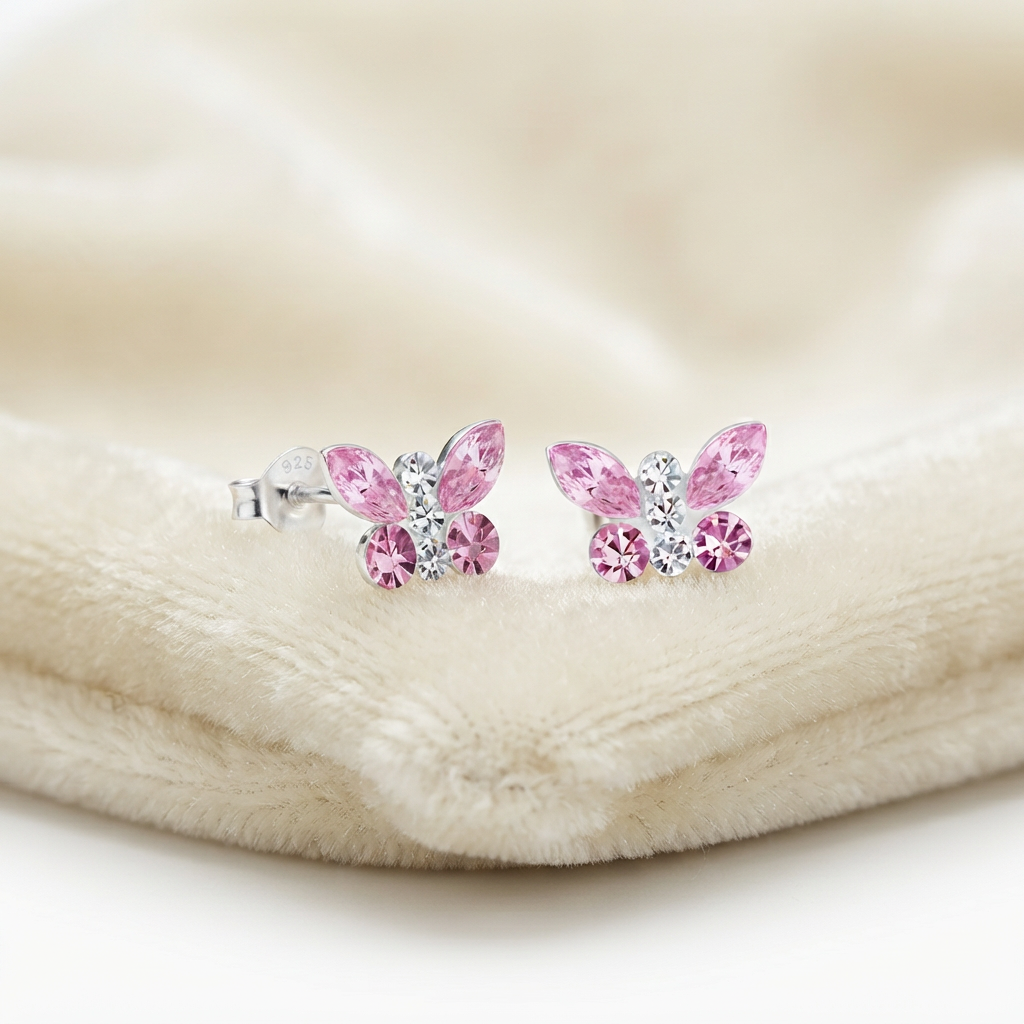 A pair of silver butterfly stud earrings with pink and clear rhinestones, resting on a soft cream-colored plush fabric.