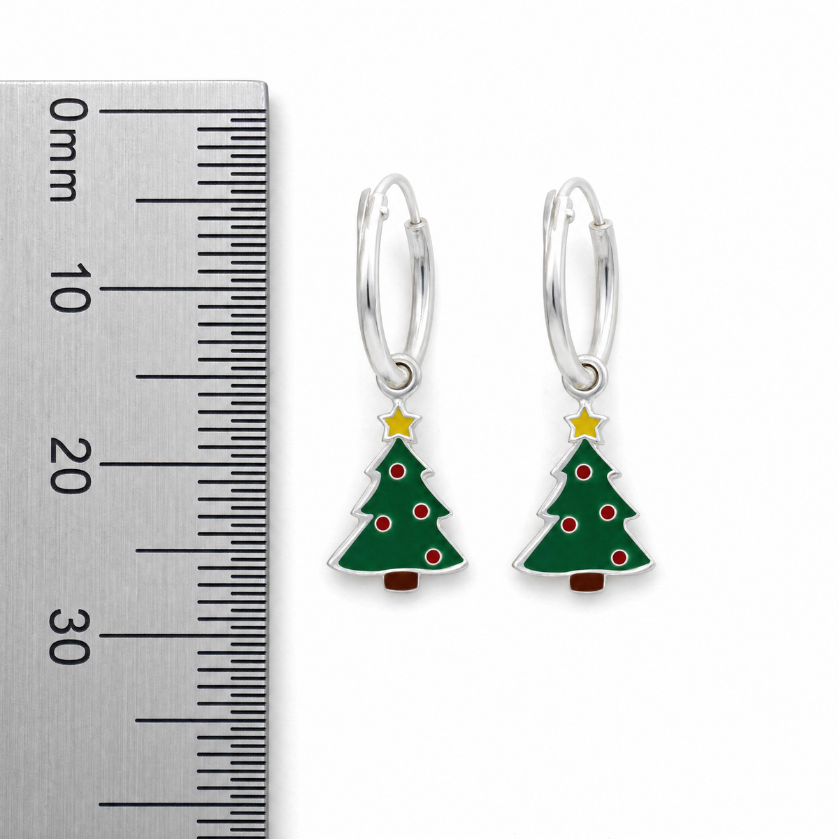 A pair of silver hoop earrings with dangling Christmas tree charms, shown next to a metal ruler for scale on a white background.