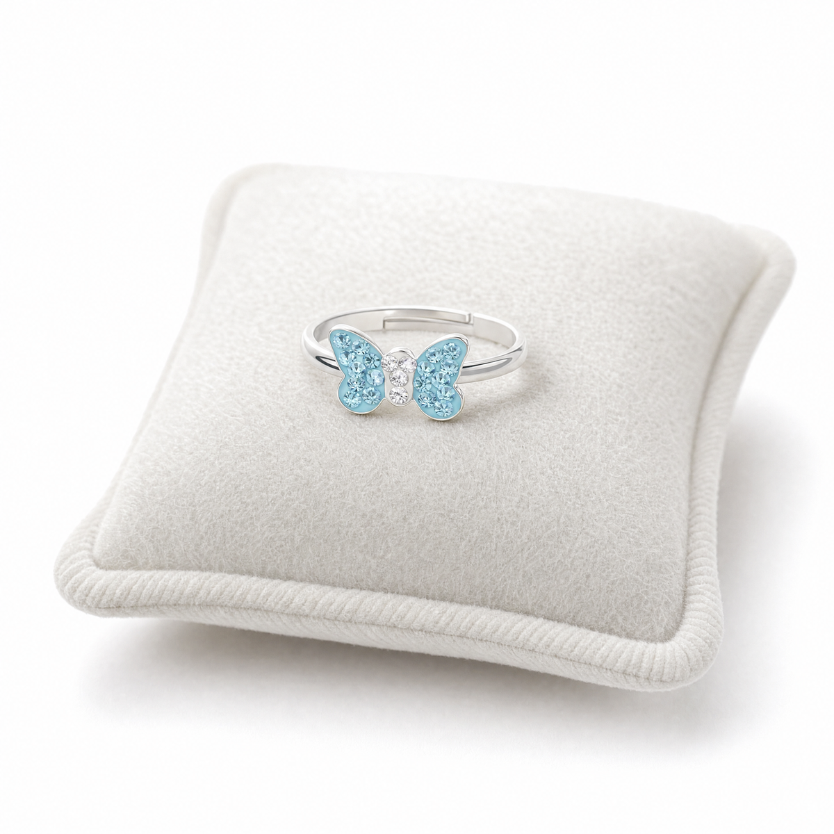 Girls silver butterfly ring with light blue gemstones and a small clear center stone, displayed on a cream-colored velvet jewelry cushion.