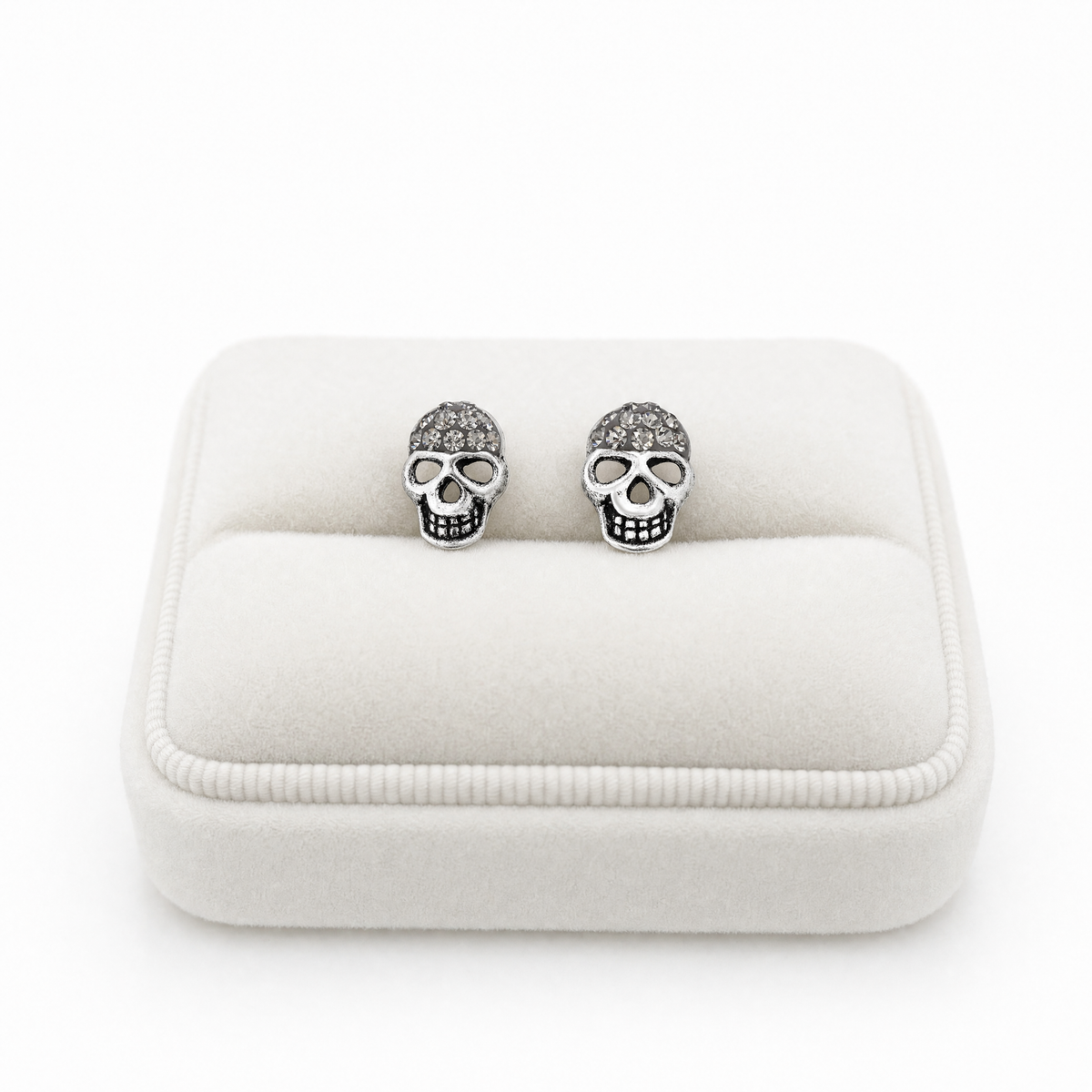 A pair of silver skull stud earrings displayed on a white velvet jewelry box against a white background.