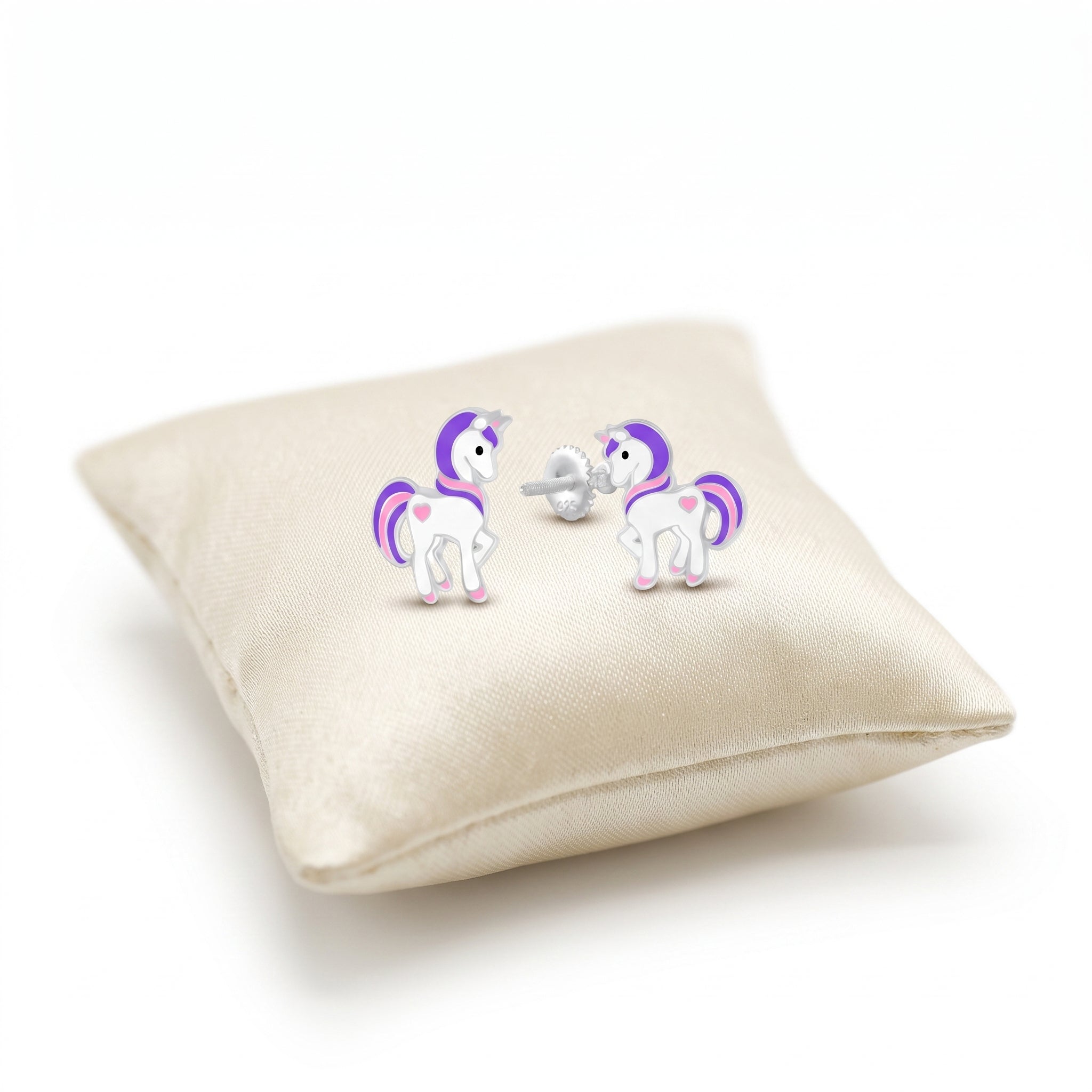 A pair of unicorn stud earrings displayed on a cream satin pillow against a white background. The earrings feature white bodies with purple and pink accents, including a purple horn, mane, tail, and small pink hearts on the side.