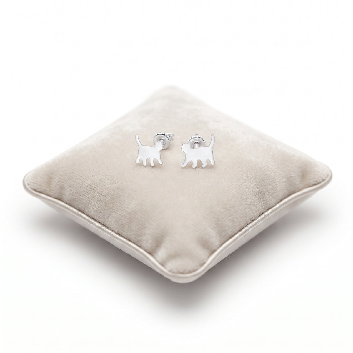 A pair of small silver cat stud earrings , displayed on a round beige velvet cushion against a white background.