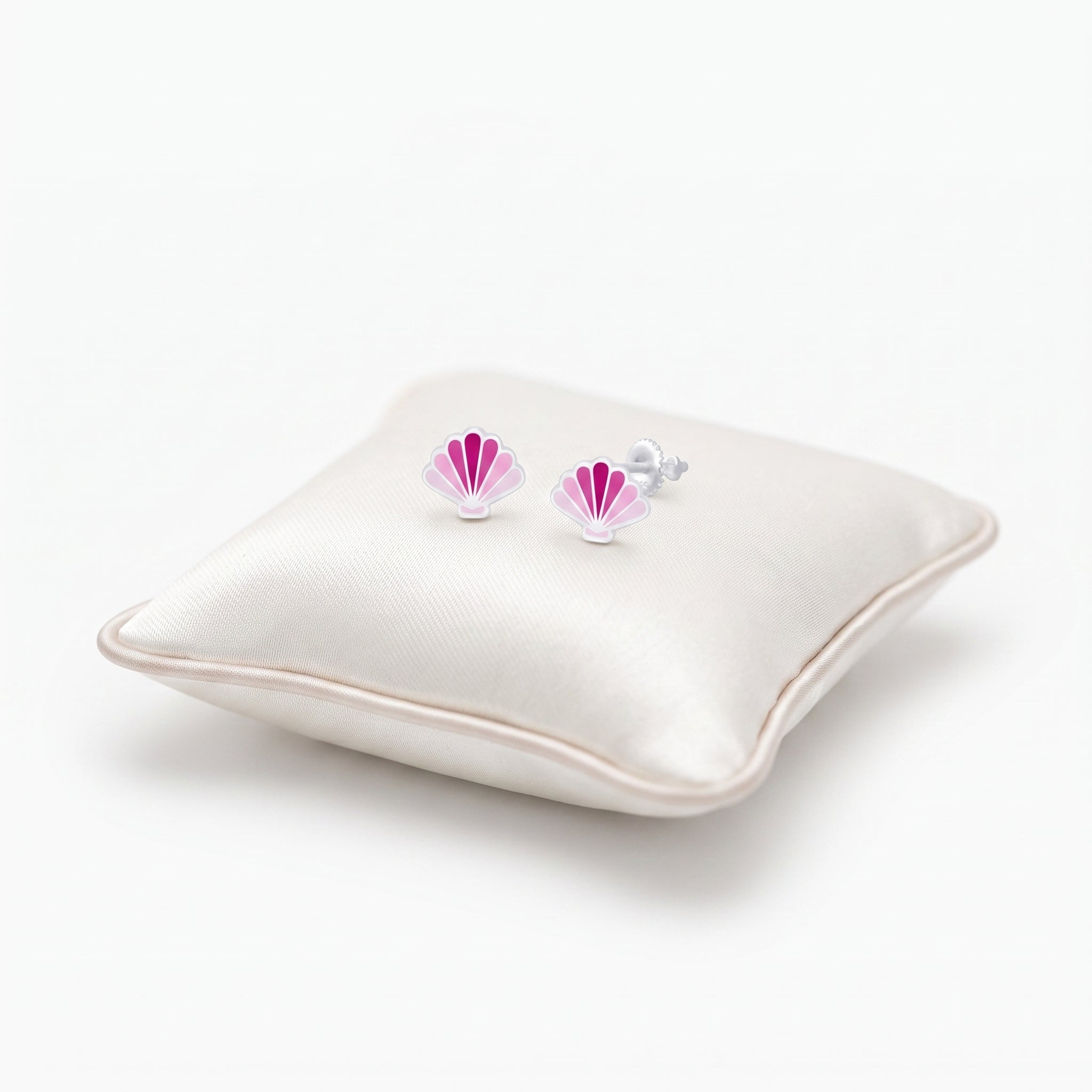 A pair of small shell-shaped stud earrings with pink and white enamel detailing and silver posts, displayed on a cream satin pillow against a white background.