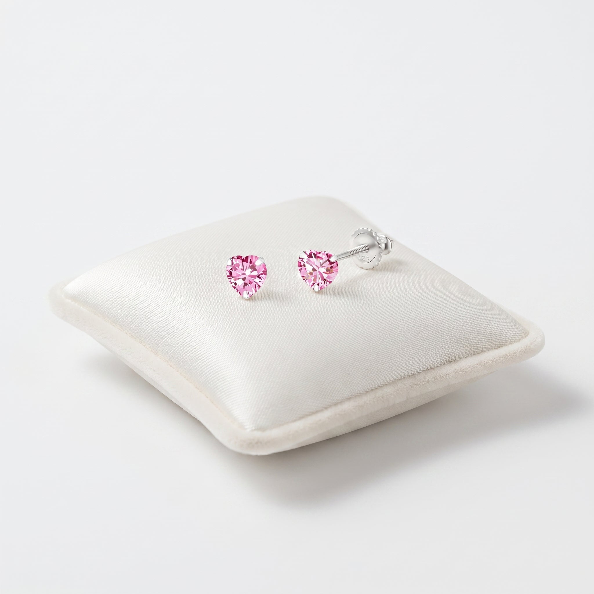 A pair of heart-shaped pink gemstone screw back stud earrings in silver settings, displayed on a cream satin jewellery pillow against a clean white background.