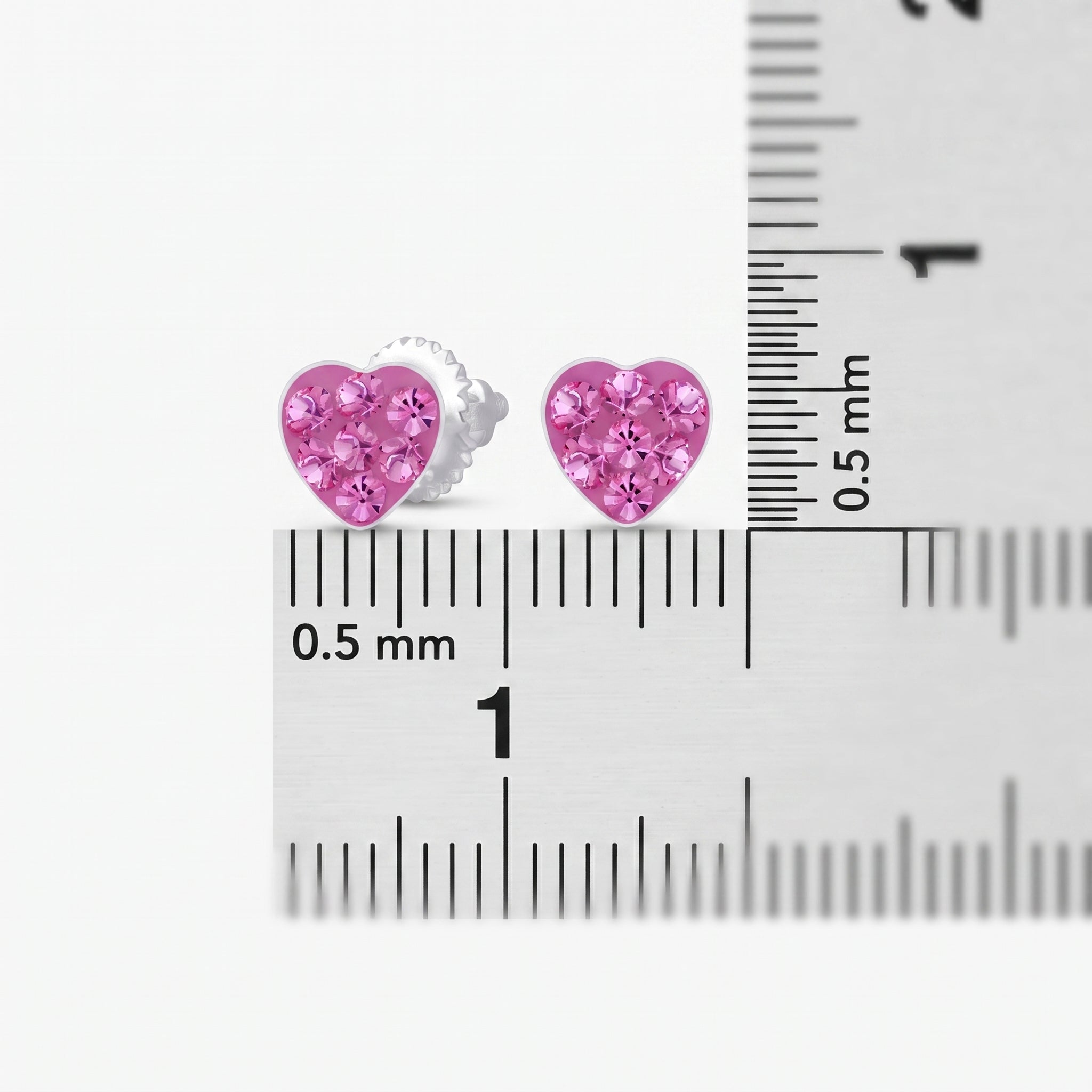 A pair of small heart-shaped stud earrings with pink rhinestones in a silver setting, shown next to a metal ruler for scale on a white background.