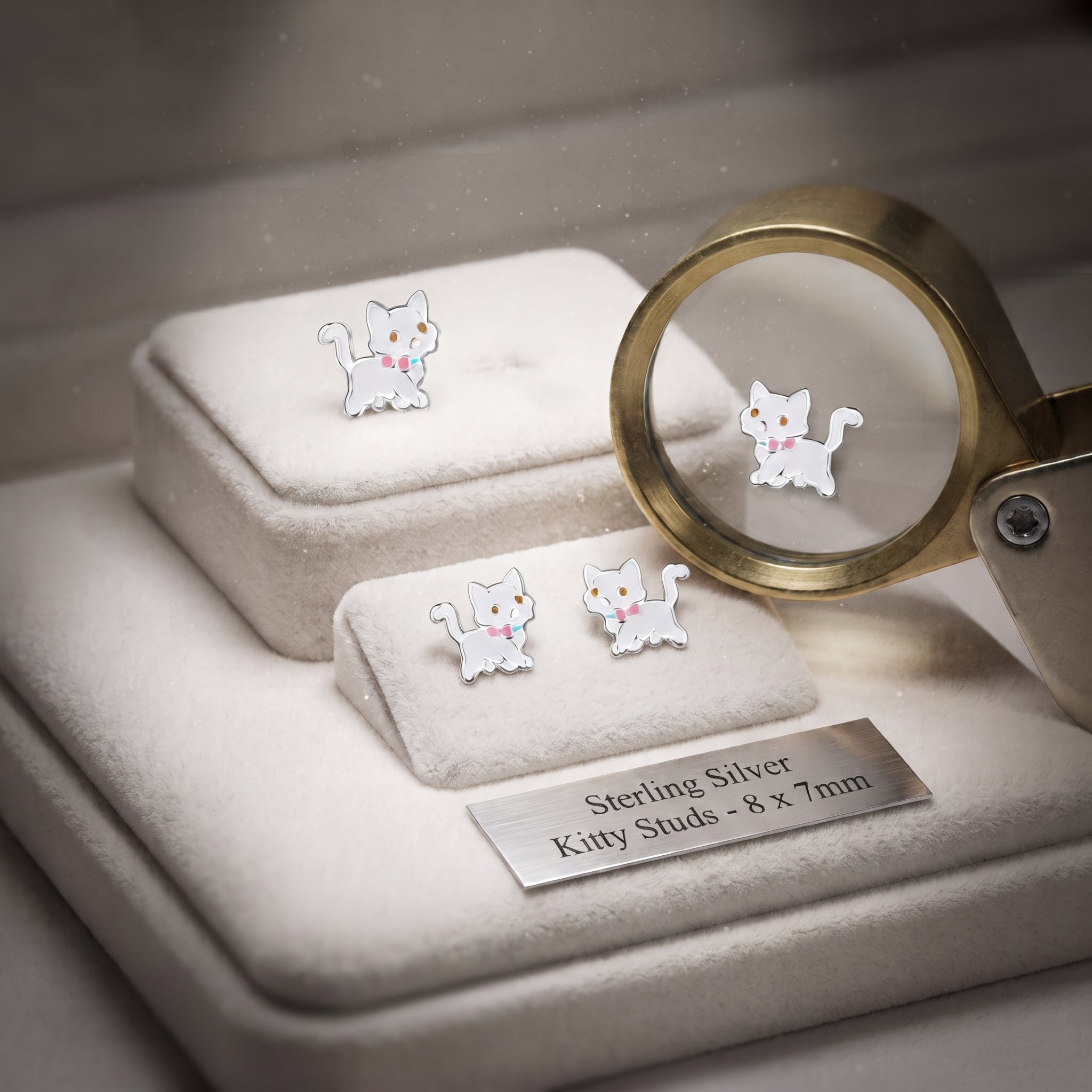 Three silver kitty stud earrings displayed on white velvet jewelry boxes, with one earring shown under a magnifying glass and a metal label reading 'Sterling Silver Kitty Studs - 8 x 7mm'.