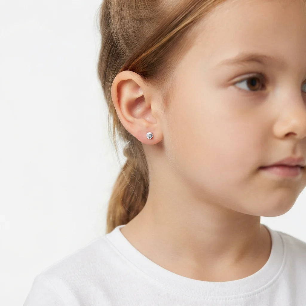 A close-up side profile of a young girl wearing a small silver stud earring with a clear gemstone, set against a clean white background.