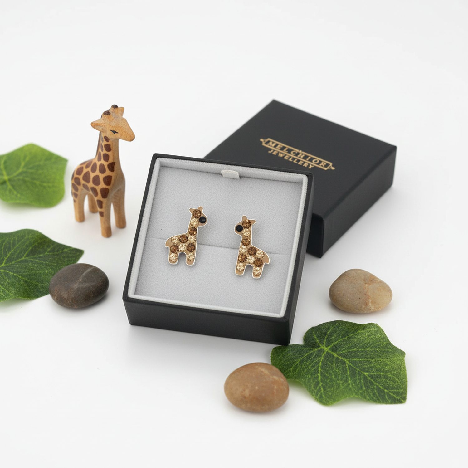 Giraffe shaped earrings for children by Melchior Jewellery displayed in a black box with decorative stones and leaves