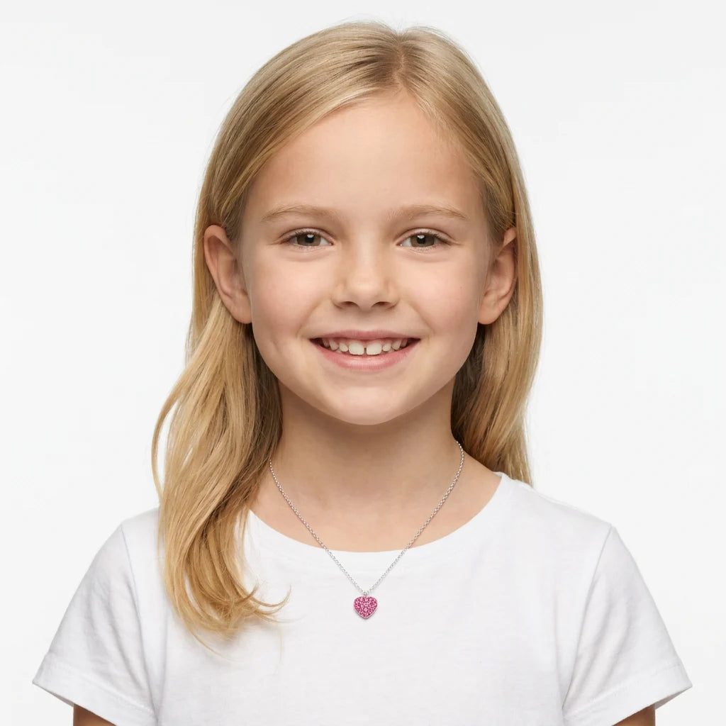 A young girl with blonde hair smiling while wearing a delicate silver chain necklace with a small pink heart pendant, set against a plain white background.