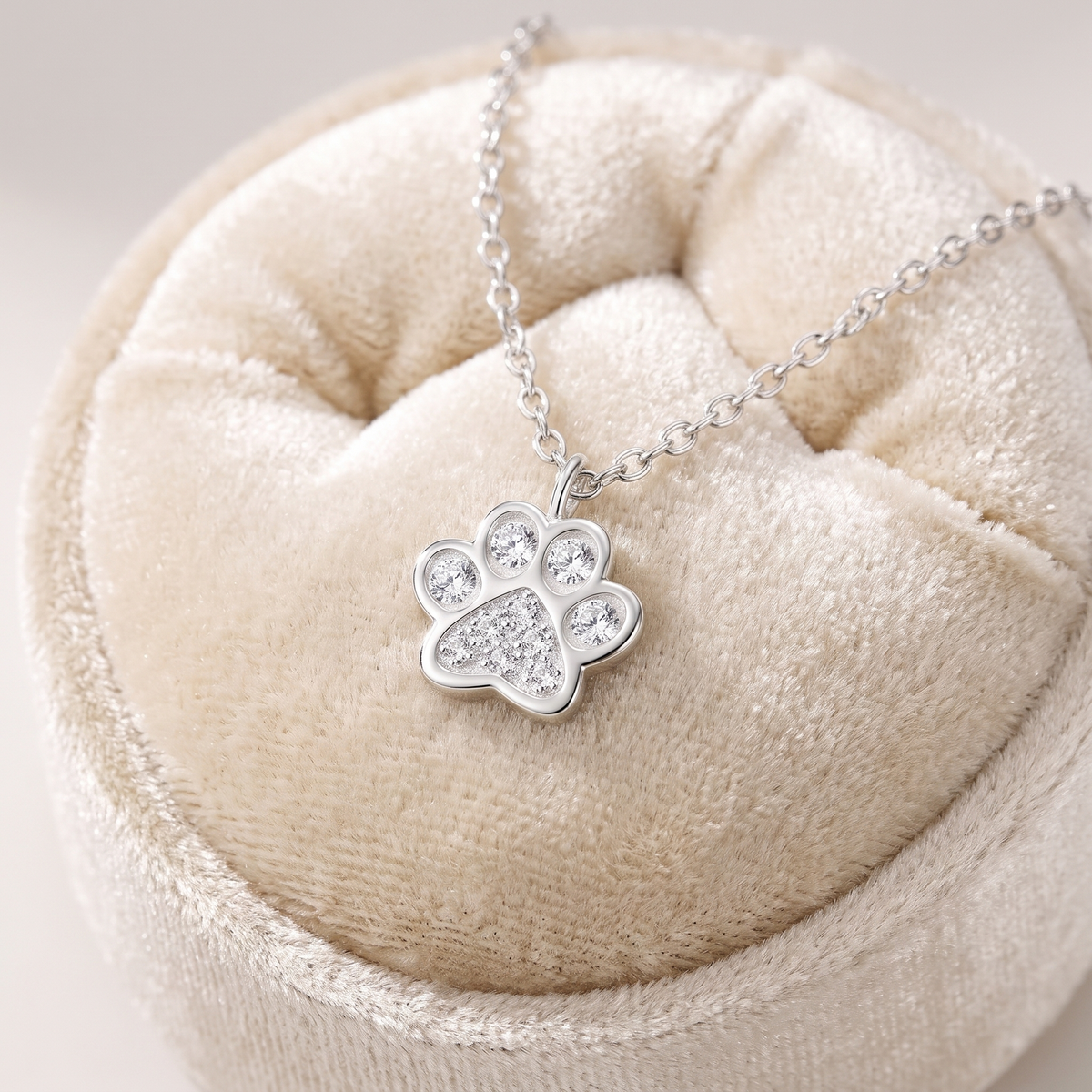 A silver paw print pendant necklace with clear crystals, displayed on a beige velvet jewelry cushion.