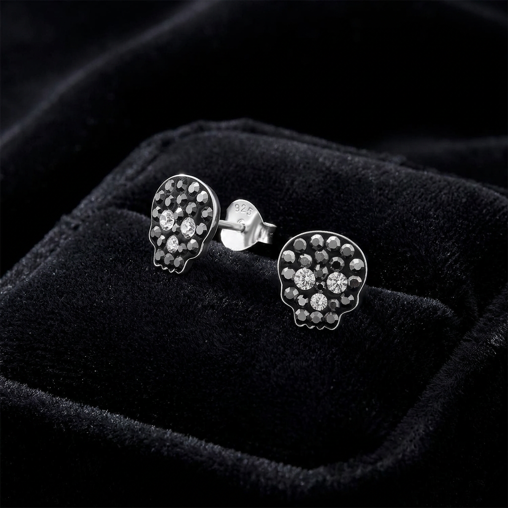 A pair of silver skull stud earrings with clear crystals, displayed in a black velvet jewelry box.