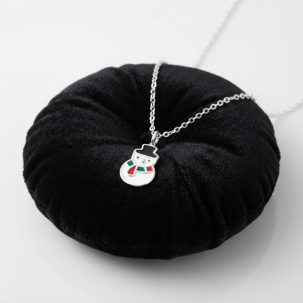 A silver snowman pendant necklace with a small red and green scarf, displayed on a black circular velvet stand against a light gray background.