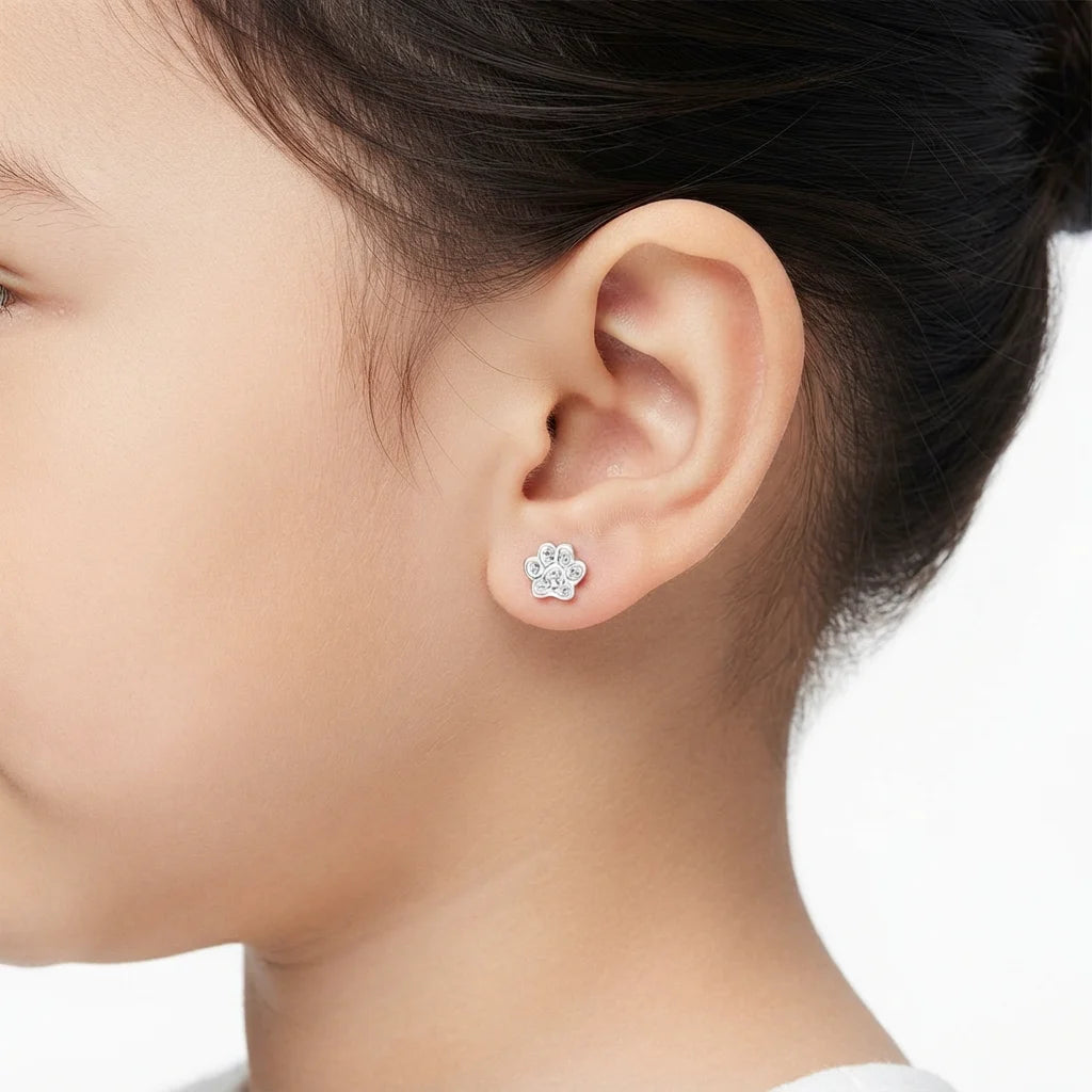 A close-up side profile of a child's head and ear wearing a small silver stud earring shaped like a flower with clear stones, against a clean white background.