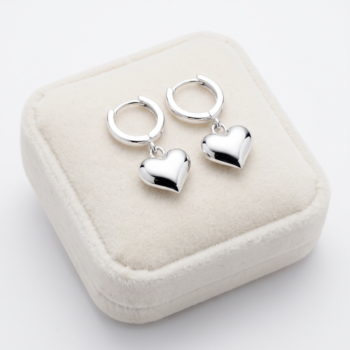 A pair of polished silver huggie hoop earrings with dangling heart charms, on Jewellery cushion