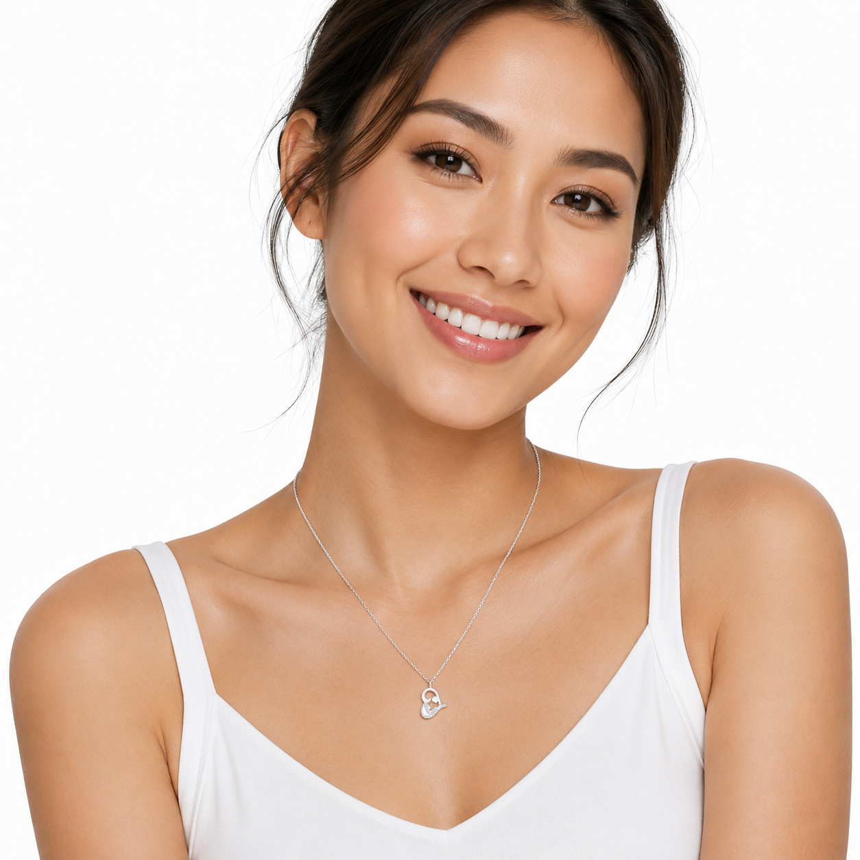 A smiling woman with dark hair pulled back wears a delicate silver necklace with a small heart-shaped pendant, paired with a white camisole top against a clean white background.