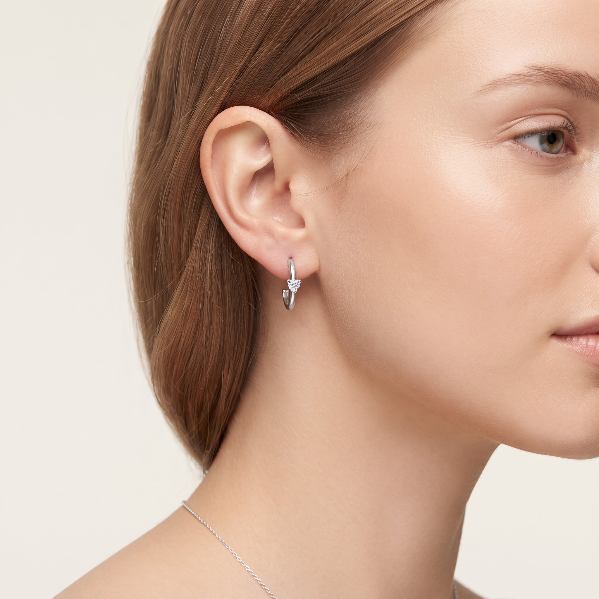 A close-up of a woman's ear and side profile wearing a small silver huggie hoop earring with a dangling silver chevron and heart-shaped crystal cluster.