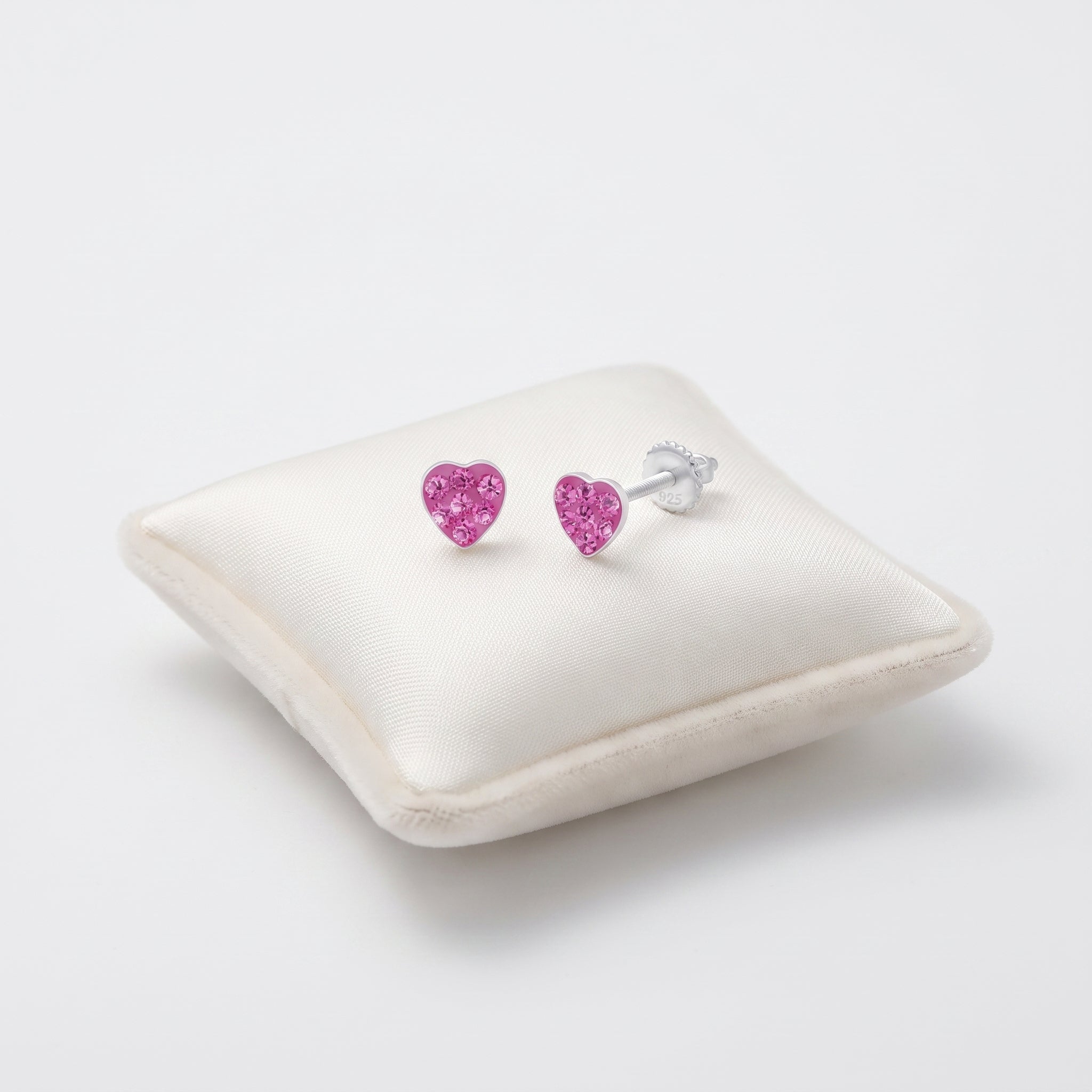 A pair of heart-shaped stud earrings with sparkling pink gemstones in silver settings, displayed on a cream satin jewelry pillow against a clean white background.