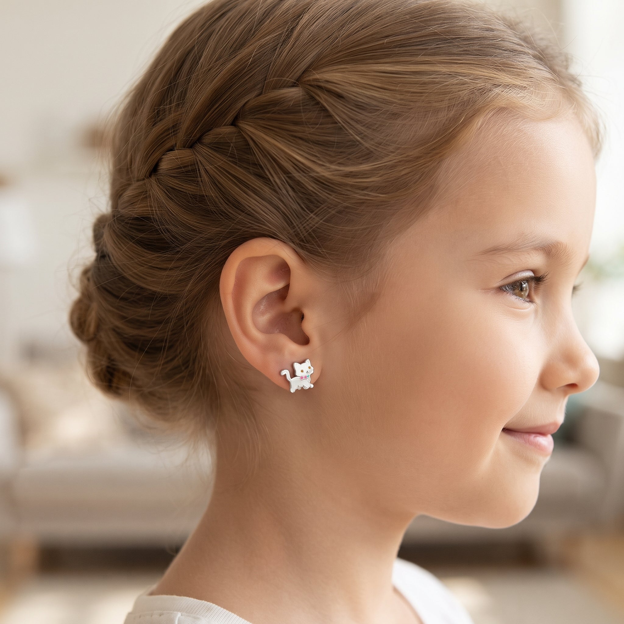 A young girl with light brown hair in a braided updo wearing a small silver stud earring shaped like a cat, shown in profile against a softly blurred indoor background.