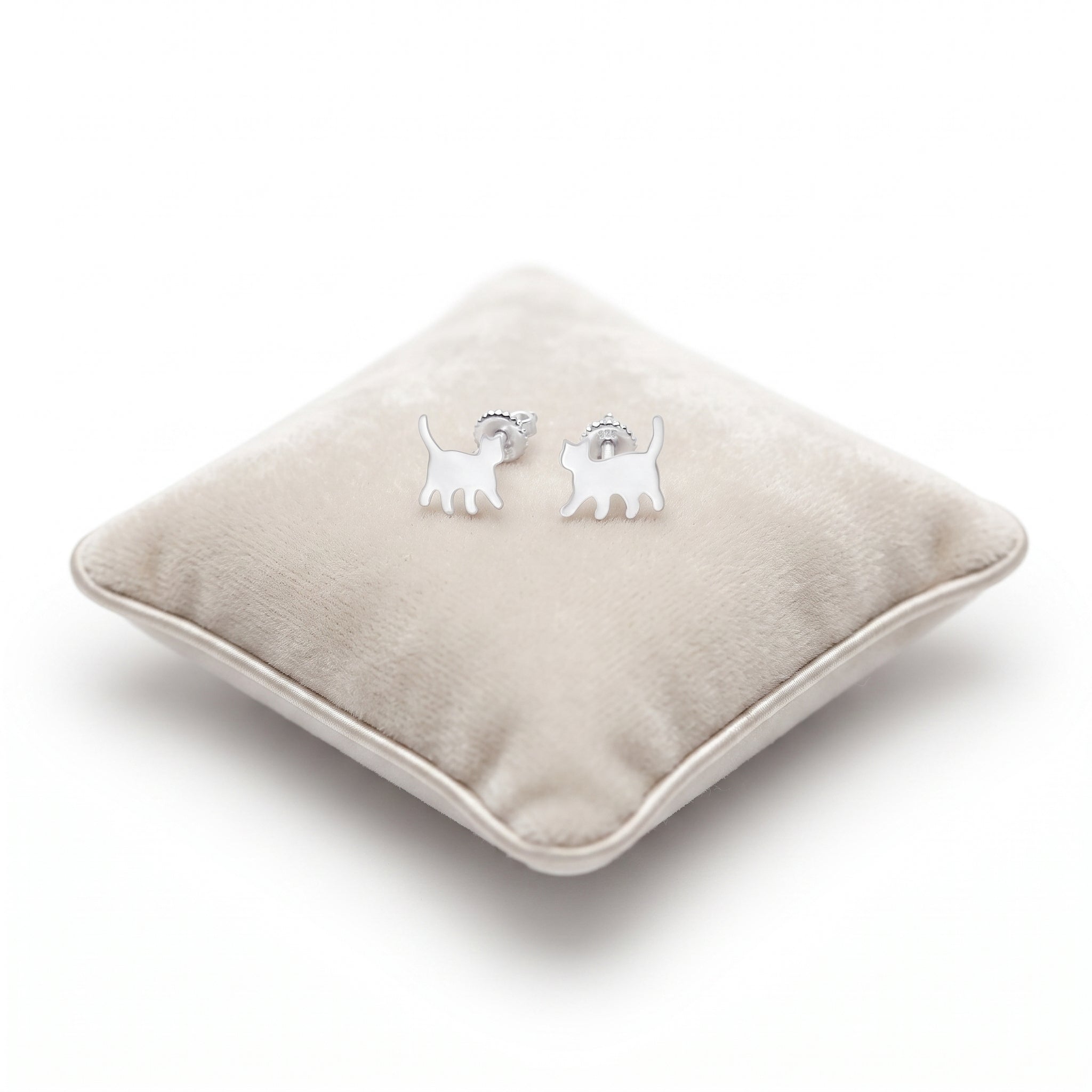 A pair of small silver cat stud earrings , displayed on a round beige velvet cushion against a white background.