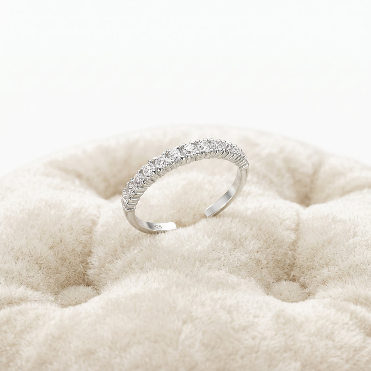 A silver-tone open band ring with a row of clear gemstones across the top, displayed on a tufted cream velvet cushion against a white background.