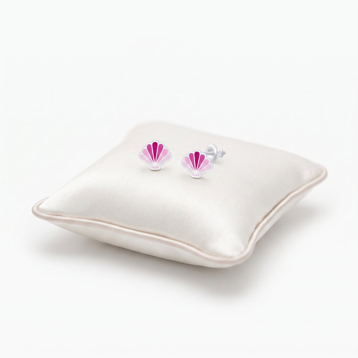 A pair of small shell-shaped stud earrings with pink and white enamel detailing and silver posts, displayed on a cream satin pillow against a white background.