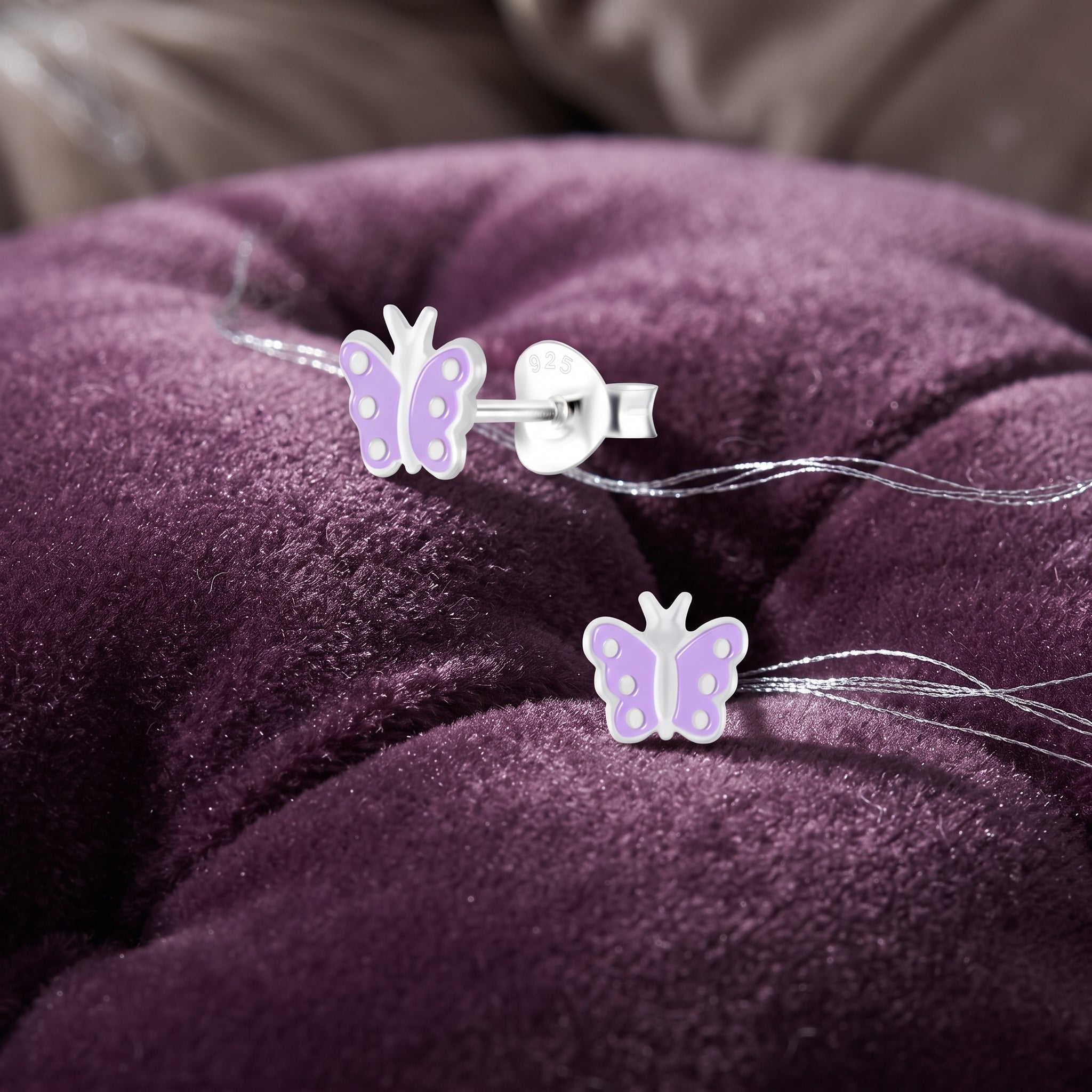 A pair of butterfly stud earrings with light purple wings and silver-toned posts, displayed on a tufted purple velvet cushion.