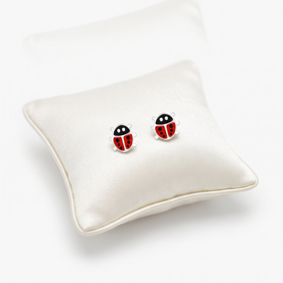 A pair of ladybug stud earrings displayed on a cream satin pillow against a clean white background.