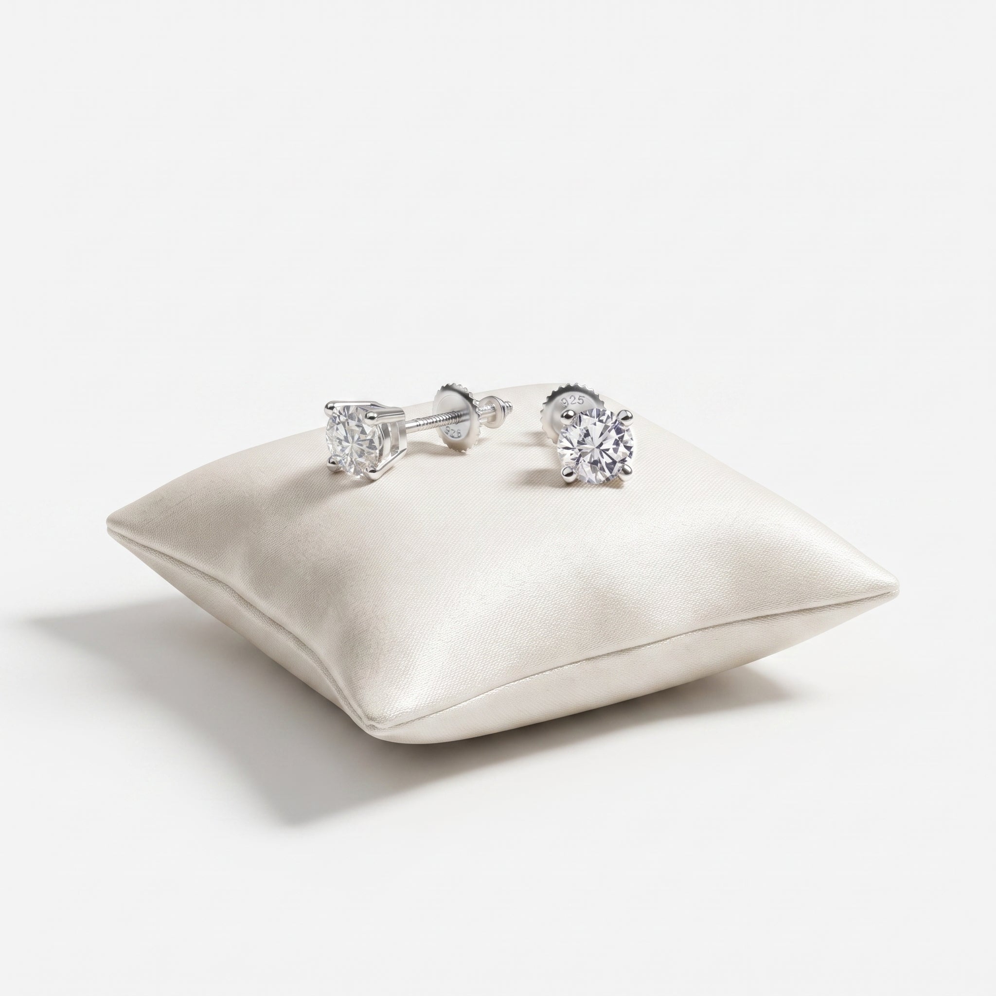 A pair of silver stud earrings with clear gemstones, displayed on a small cream satin cushion against a clean white background.