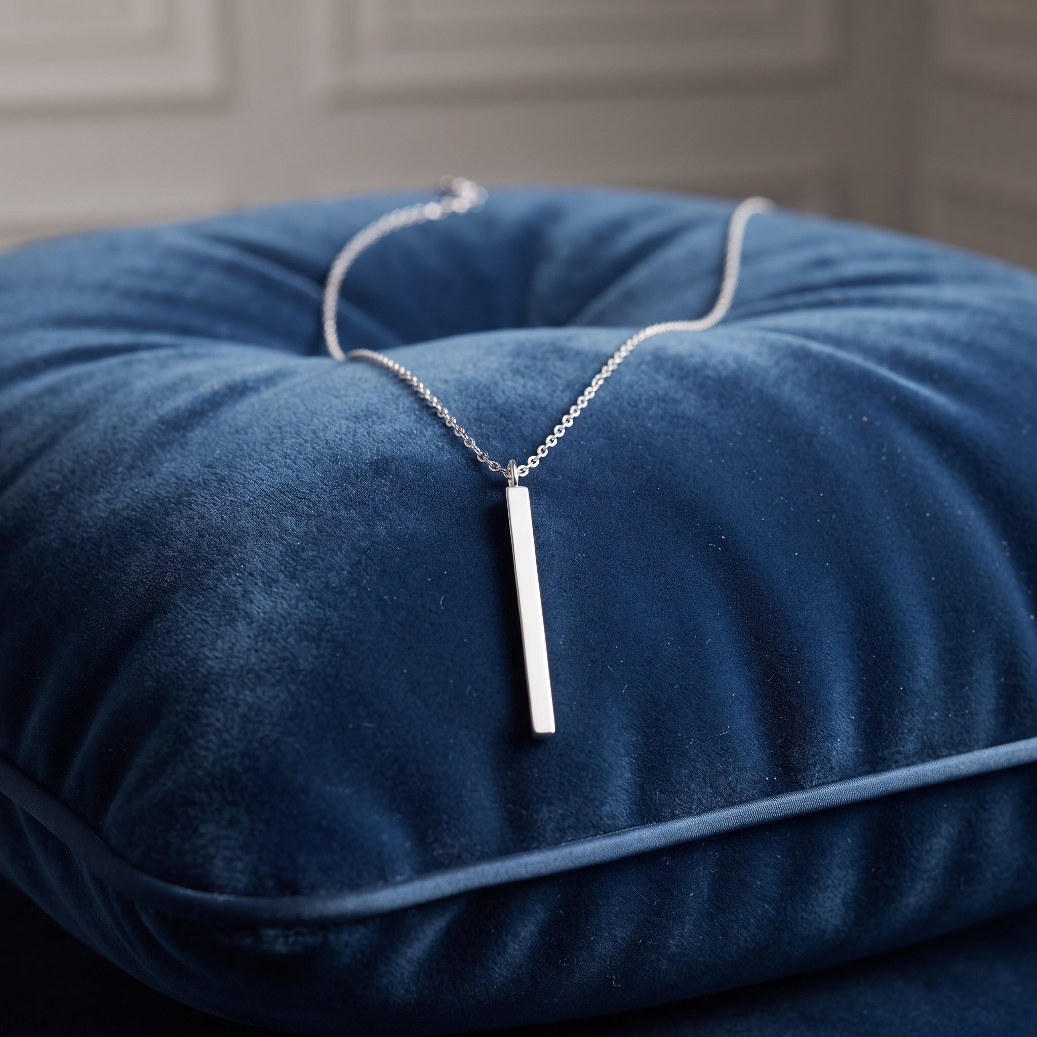 A silver bar necklace with a delicate chain, displayed on a plush navy blue velvet cushion.