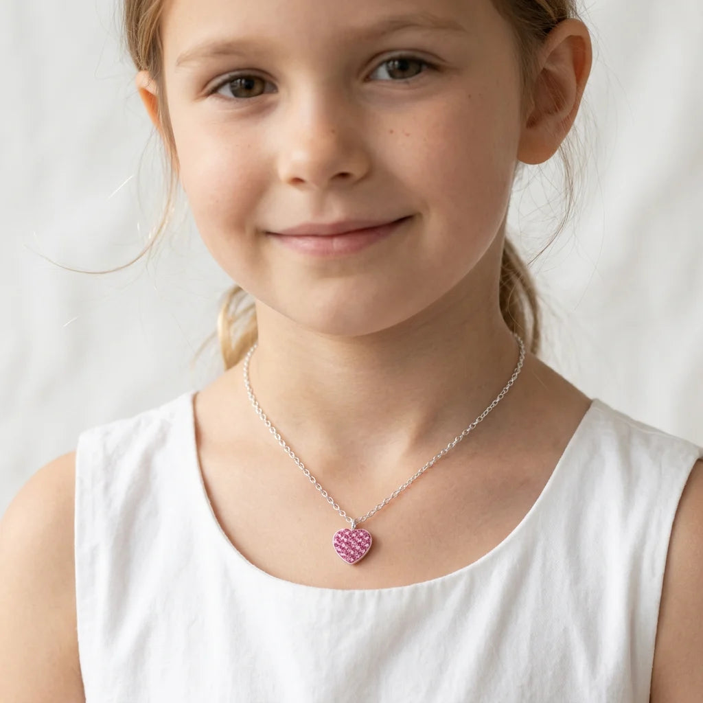 A young girl with light brown hair tied back wears a silver chain necklace with a pink heart-shaped charm, paired with a white sleeveless top against a soft white background.