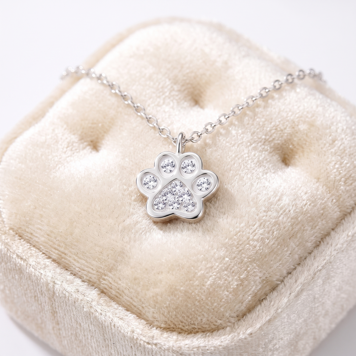A silver paw print pendant necklace with clear rhinestones, displayed on a cream-colored velvet jewelry box.