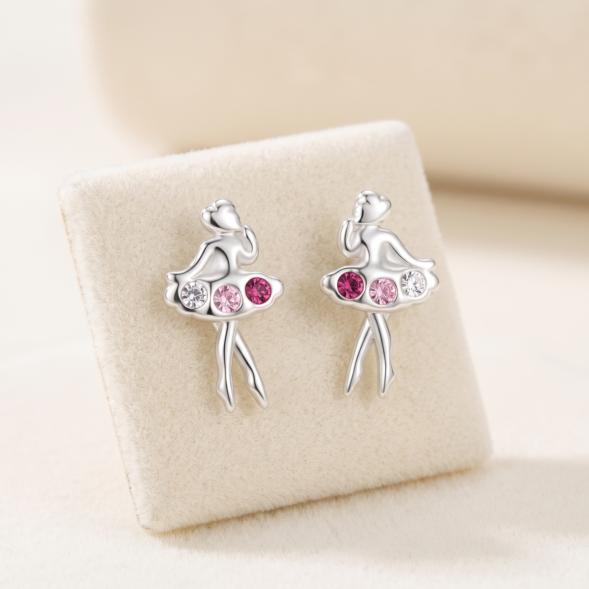 A pair of silver stud earrings shaped like ballerinas, featuring pink and clear rhinestones, displayed on a cream-colored jewelry stand.
