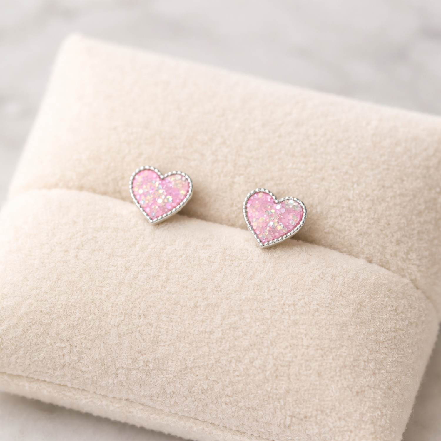 Pink glitter heart stud earrings for kids by Melchior Jewellery displayed on a soft beige cushion