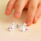 Sterling silver rabbit earrings for kids with pink heart details by Melchior Jewellery next to a child's fingers